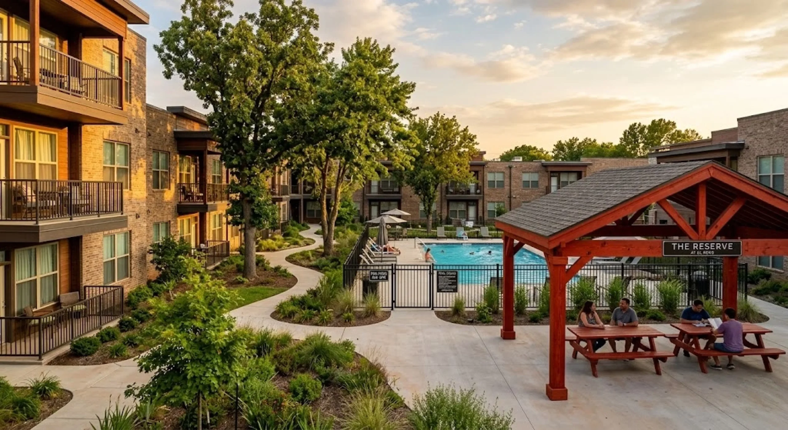 Wide-angle, golden-hour photograph of The Reserve at El Reno apartment courtyard, featuring a rustic wooden picnic pergola, people seated at a picnic table, a large swimming pool behind a black fence, mature trees, and multi-story brick apartment buildings with balconies in the background.