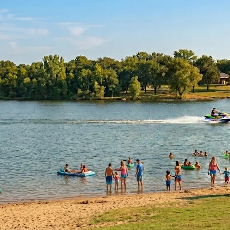 View of a busy lake with families on the beach and in the water, people floating, a jet ski rider, and kayaks. A historic stone church is visible on the far hill.